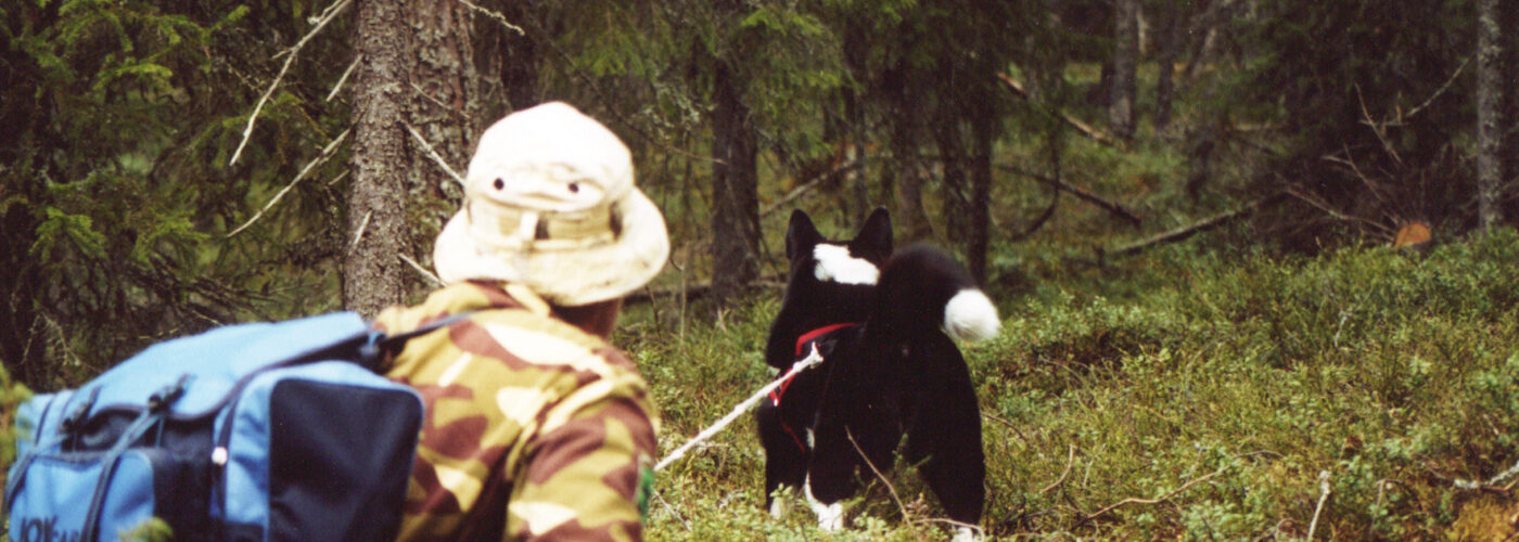 Eräasuinen henkilö kuusimetsässä mustavalkoisen koiran kanssa.
