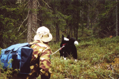 Eräasuinen henkilö kuusimetsässä mustavalkoisen koiran kanssa.